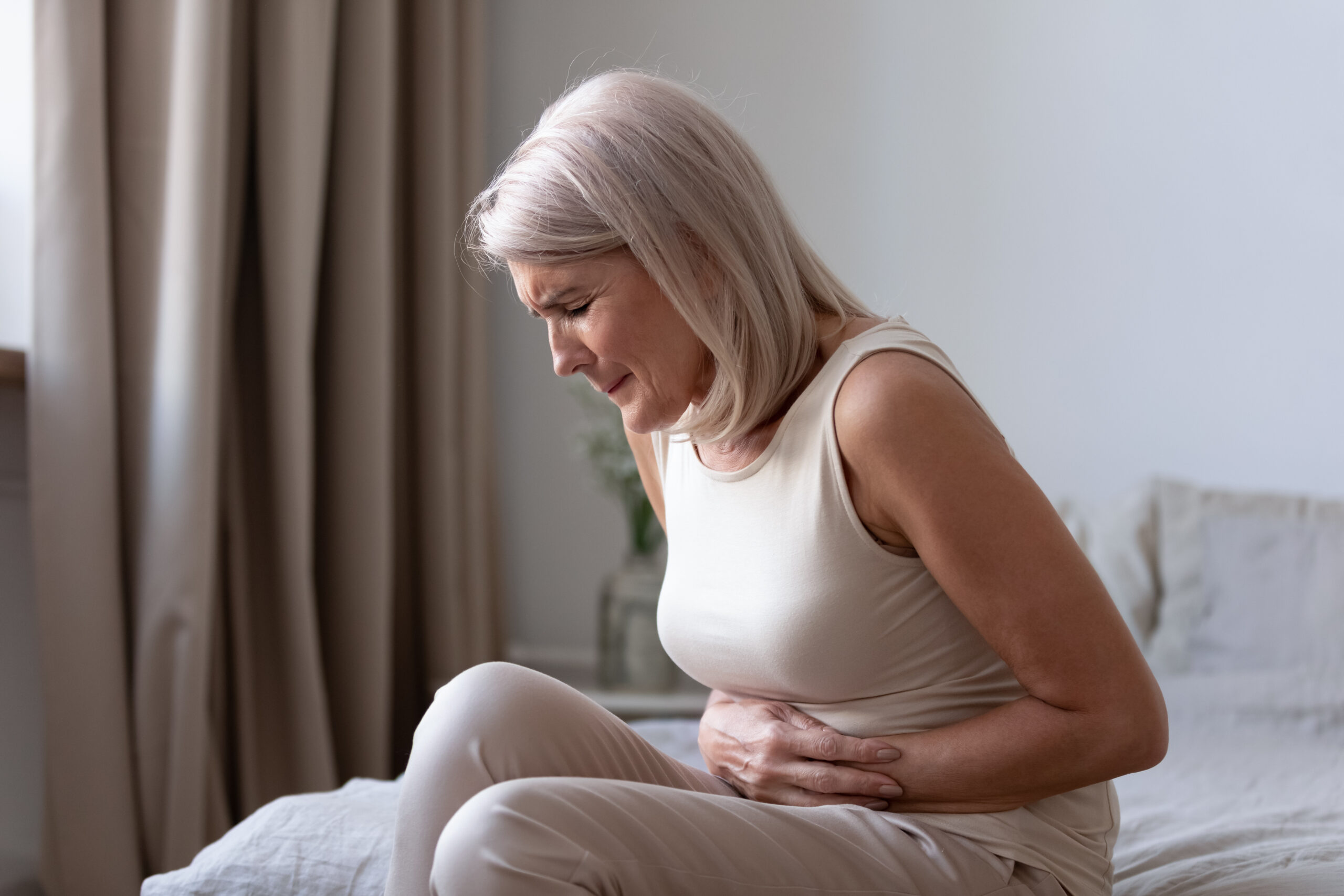 Middle-aged woman on bed wincing in abdominal pain, potential gastrointestinal issue.