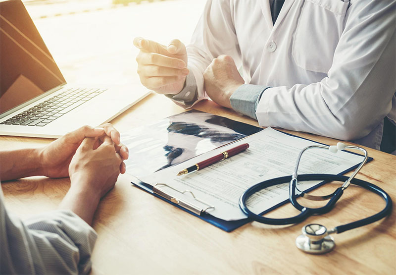 Doctor discusses patient care in medical office with forms and laptop on desk.